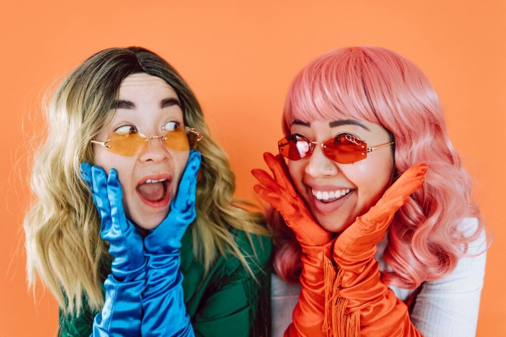 Two joyful women in colorful wigs and gloves expressing surprise on an orange background.