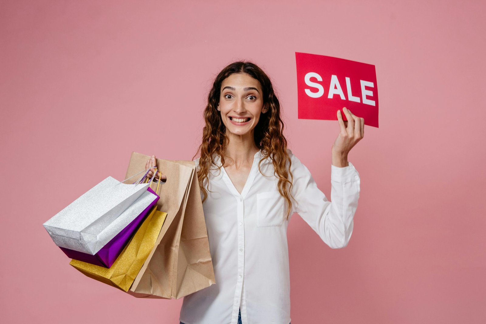 Joyful woman holding shopping bags and a sale sign, ready for a shopping spree on a pink background.